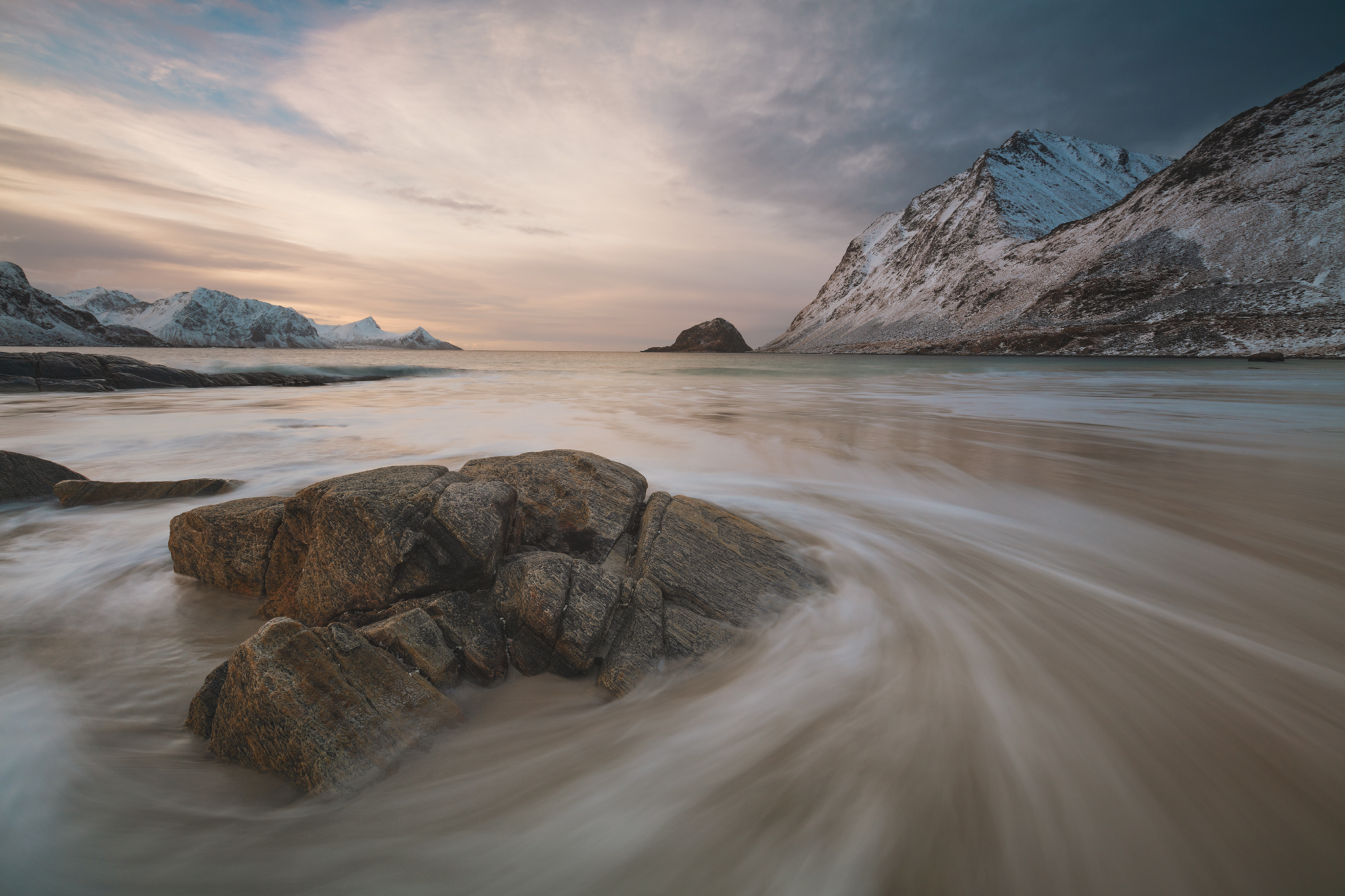 Long exposure of waves circling a rock on a mountainous background in the Lofoten Islands of Norway